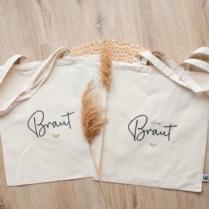 Op de afbeelding: Twee off-white katoenen draagtassen met hengsels. De linker tas heeft de zwarte cursieve tekst Braut met een klein goudkleurig hartje eronder. De rechter tas heeft de zwarte cursieve tekst Team Braut met een klein goudkleurig hartje. Gedroogd gras.