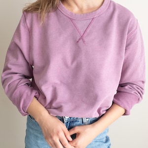 以下が含まれることがあります： 丸首と長袖のモーヴ色のスウェットシャツ。ネックラインにはV字型のステッチディテールがあります。袖口と裾は濃いモーヴ色です。スウェットシャツは、ライトブルーのデニムジーンズと組み合わせています。
