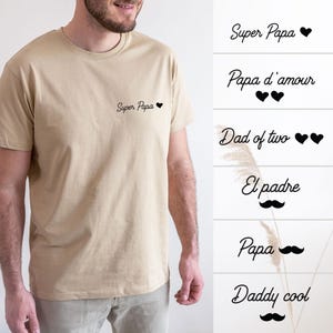 Pode incluir: Uma camiseta bege com gola redonda com o texto "Super Papa" e um pequeno coração preto no peito. Opções de texto adicionais incluem "Papa d'amour", "Dad of two", "El padre", "Papa" e "Daddy cool".