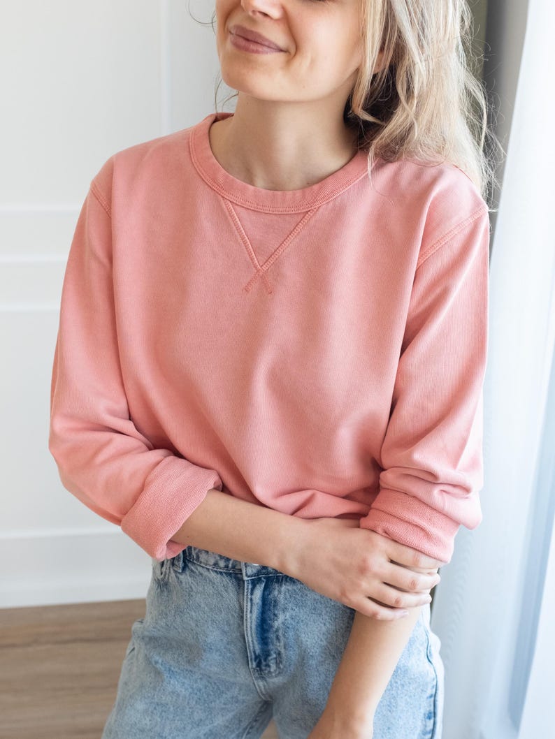Puede incluir: Una sudadera de color coral con cuello redondo y mangas largas. La sudadera tiene un corte holgado y se combina con vaqueros de mezclilla de lavado claro. Los pu&ntilde;os y el cuello tienen una textura acanalada.