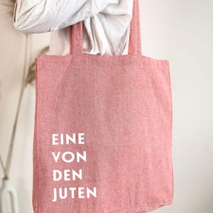 May include: A red tote bag with the words "EINE VON DEN JUTEN" printed in white. The bag is made of a textured fabric and has long straps for carrying over the shoulder. The bag is being held by a person wearing a white shirt and blue jeans.