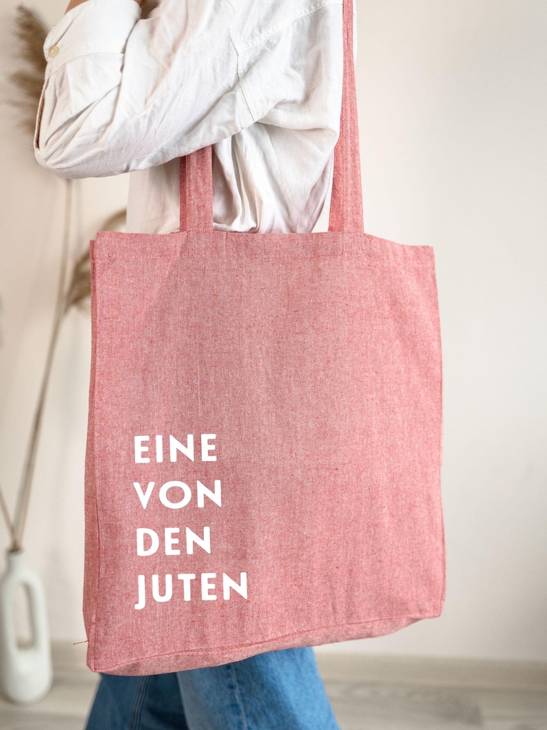 May include: A red tote bag with the words "EINE VON DEN JUTEN" printed in white. The bag is made of a textured fabric and has long straps for carrying over the shoulder. The bag is being held by a person wearing a white shirt and blue jeans.