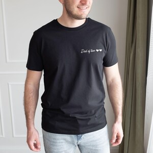May include: Black crew neck t-shirt with the text "Dad of two" and two white hearts. The shirt is a solid black color and has short sleeves. The shirt is made of a soft material.