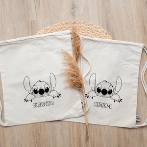 Peut inclure: Deux sacs à cordon de serrage blanc cassé avec une illustration en noir et blanc de Stitch de Lilo & Stitch. Un sac est étiqueté "Christina" et l'autre "Michael". Les sacs ont des cordons blancs et sont posés sur une surface en bois clair.