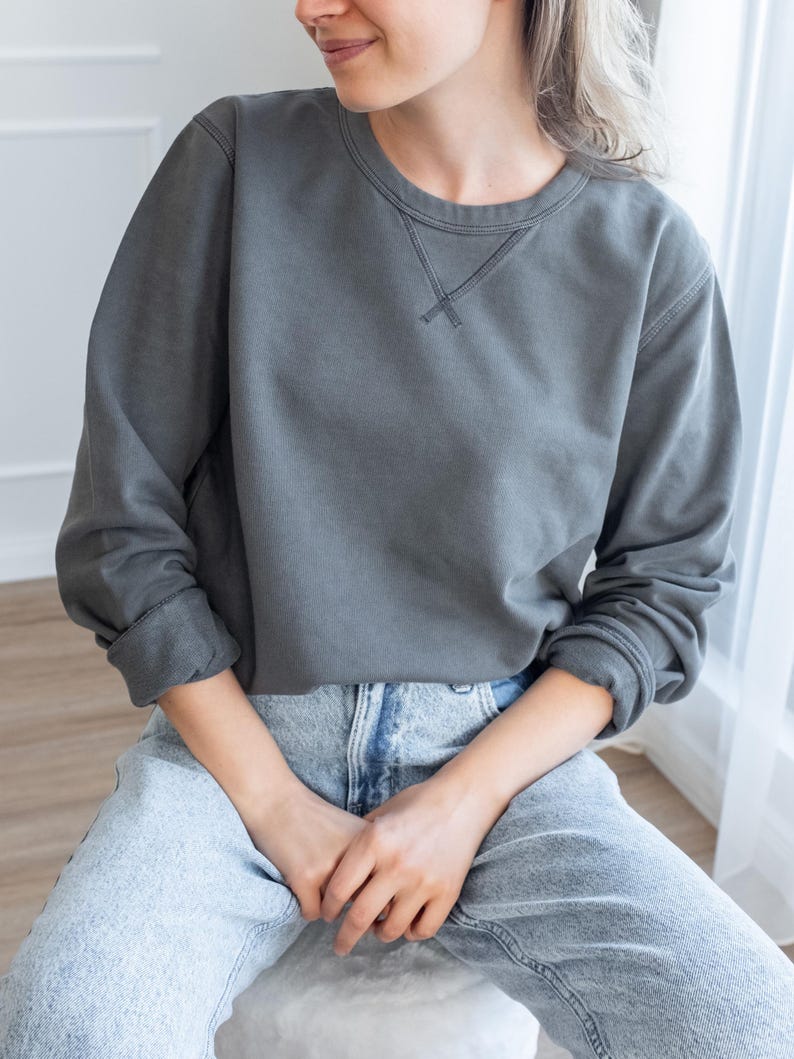 Puede incluir: Una sudadera gris carb&oacute;n con cuello redondo y mangas largas. La sudadera tiene un detalle cosido en el cuello y se combina con vaqueros de mezclilla de lavado claro. La modelo est&aacute; sentada, con las manos entrelazadas en el regazo.