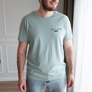 May include: A light green t-shirt with the text "Papa d'amour" and two black hearts. The shirt is short-sleeved and made of a soft material. The model is wearing blue jeans.