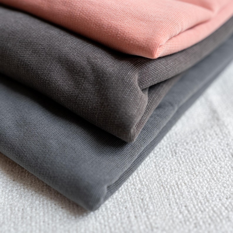 Puede incluir: Primer plano de tela doblada en tonos grises y un color rosa empolvado. La tela est&aacute; apilada en capas, con la tela rosa en la parte superior. La textura de la tela es visible, lo que sugiere un material suave.