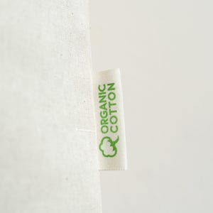 May include: Close-up of a white fabric with a tag that reads "ORGANIC COTTON" in green letters. A green cotton plant icon is below the text. The fabric has a textured appearance.