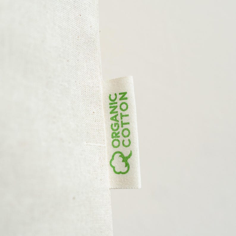 May include: Close-up of a white fabric with a tag that reads "ORGANIC COTTON" in green letters. A green cotton plant icon is below the text. The fabric has a textured appearance.