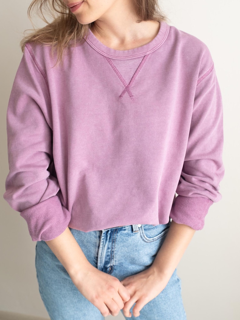 Puede incluir: Una sudadera de color malva con cuello redondo y mangas largas. El escote presenta un detalle decorativo de punto de cruz. Los pu&ntilde;os y el dobladillo son de un tono malva m&aacute;s oscuro. La sudadera se combina con unos vaqueros azules.