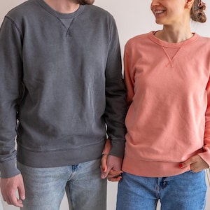 Puede incluir: Dos personas con sudaderas de cuello redondo a juego. Una sudadera es de color gris carb&oacute;n descolorido, la otra es de color coral. Ambas tienen un escote cosido y se combinan con vaqueros azules. La pareja se da la mano.