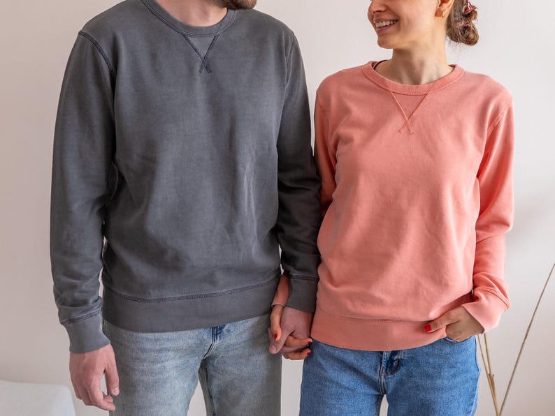 Puede incluir: Dos personas con sudaderas de cuello redondo a juego. Una sudadera es de color gris carb&oacute;n descolorido, la otra es de color coral. Ambas tienen un escote cosido y se combinan con vaqueros azules. La pareja se da la mano.