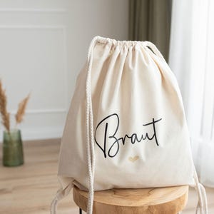 Puede incluir: Una bolsa de cordón blanca con la palabra "Braut" escrita en letra negra y un corazón dorado. La bolsa está sentada en un taburete de madera.