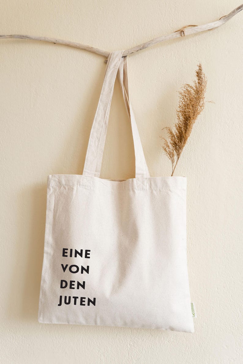May include: A cream-colored tote bag with black text that reads "EINE VON DEN JUTEN." The bag hangs from a wooden branch, with a dried plant stem placed inside. The bag is made of a sturdy fabric.