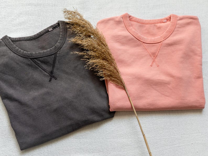Puede incluir: Se muestran dos sudaderas de cuello redondo, una gris antracita y otra rosa coral. Ambas camisas tienen un detalle de cuello en V cosido. Un tallo de planta seca descansa sobre las camisas. Las camisas est&aacute;n cuidadosamente dobladas.