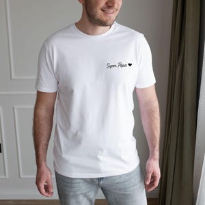 May include: White t-shirt with the text "Super Papa" and a small black heart. The shirt is a classic crew neck style and appears to be made of a soft, comfortable material. The text is in a handwritten style.