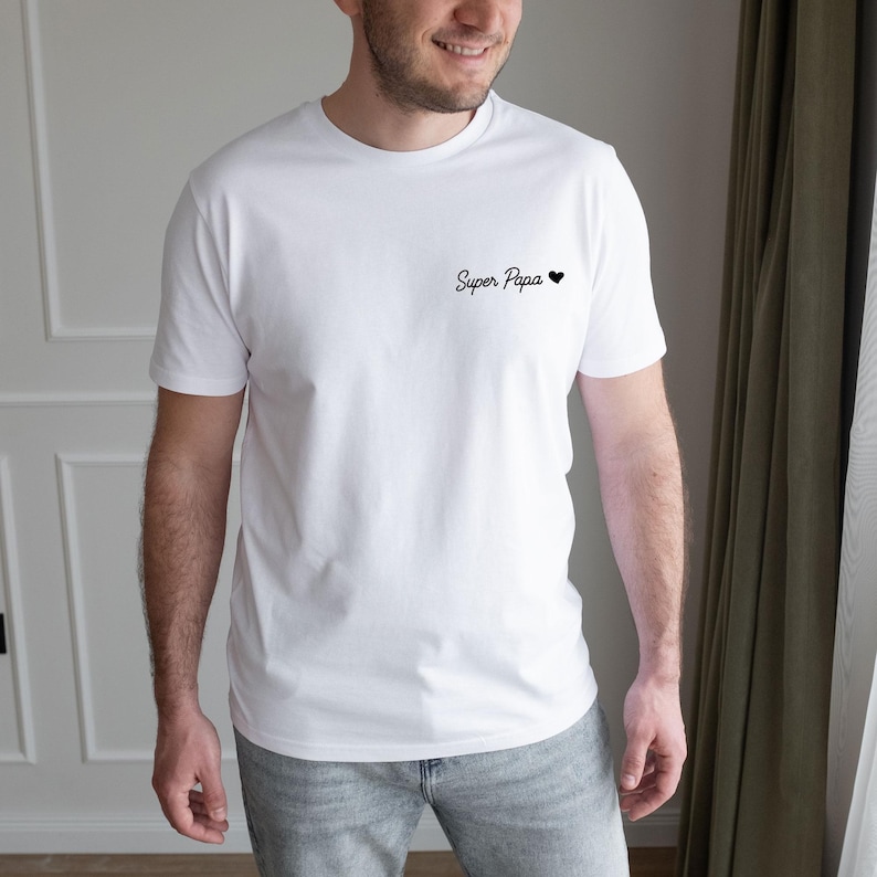 May include: White t-shirt with the text "Super Papa" and a small black heart. The shirt is a classic crew neck style and appears to be made of a soft, comfortable material. The text is in a handwritten style.