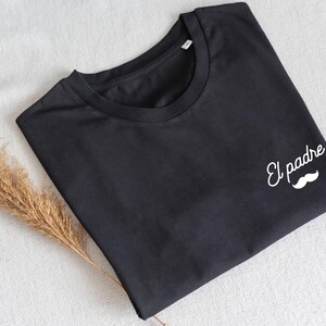 May include: Black t-shirt with the words "El padre" and a white mustache graphic. The shirt is folded and lies on a white surface, with a dried plant stem to the side. A casual apparel item.