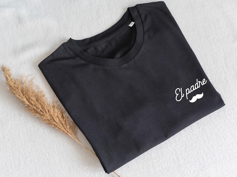 May include: Black t-shirt with the words "El padre" and a white mustache graphic. The shirt is folded and lies on a white surface, with a dried plant stem to the side. A casual apparel item.