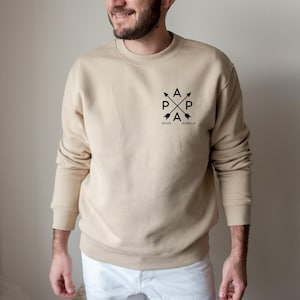 Puede incluir: Sudadera beige de cuello redondo con un diseño gráfico negro. El diseño presenta flechas cruzadas con las letras "A" y "P" y los nombres "MARK" e "ISABELA" debajo. La sudadera es de color neutro.