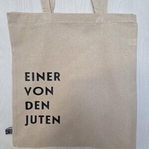 May include: Beige tote bag with the text "EINER VON DEN JUTEN" in black. The bag has two handles and a small label on the bottom left corner. The bag is made of a natural, woven material.