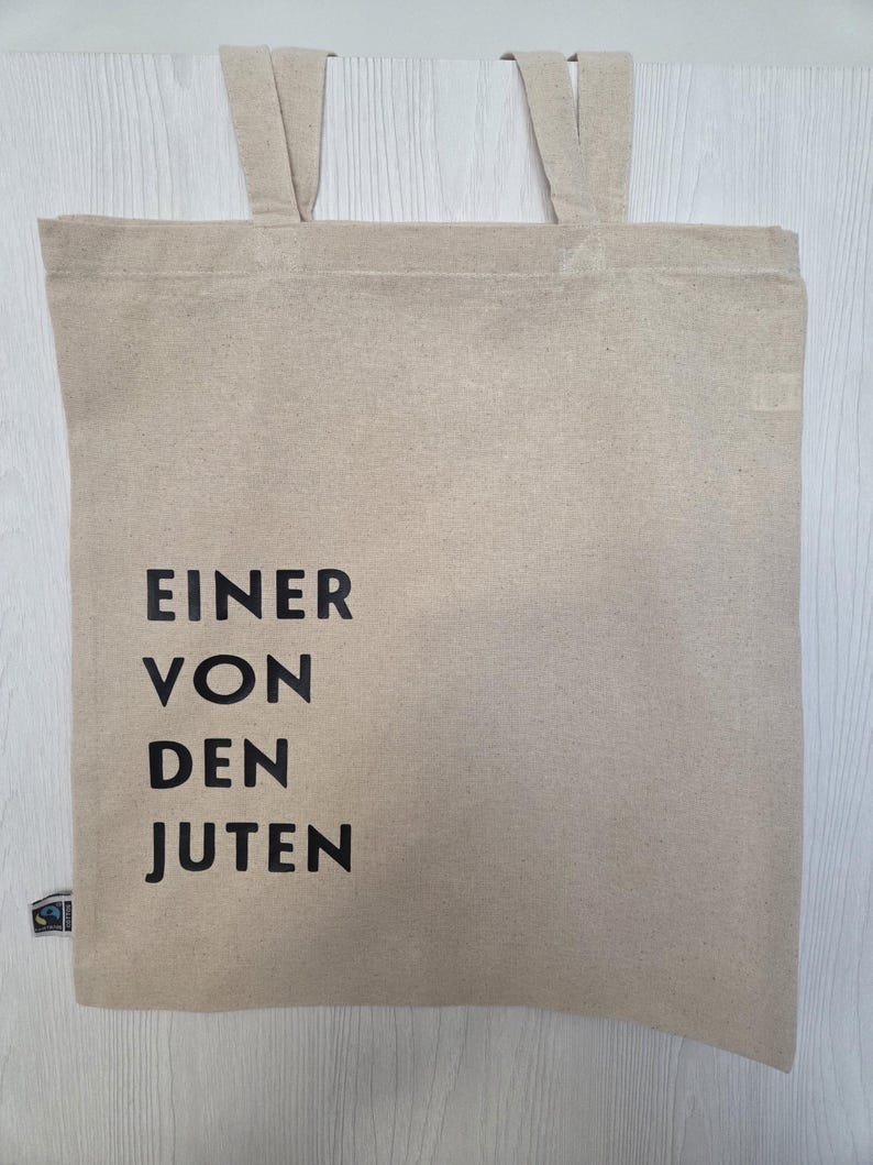 May include: Beige tote bag with the text "EINER VON DEN JUTEN" in black. The bag has two handles and a small label on the bottom left corner. The bag is made of a natural, woven material.