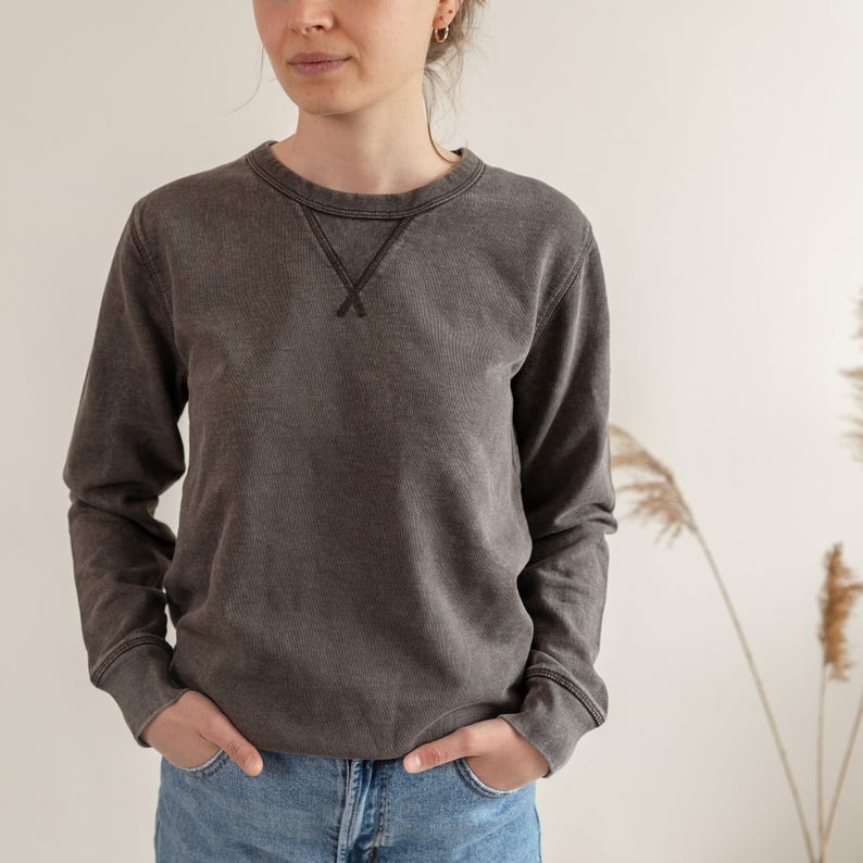 Puede incluir: Una sudadera marr&oacute;n oscuro con cuello redondo y mangas largas. El escote presenta un detalle de costura en forma de V. La sudadera se usa con vaqueros azules. El estilo general es casual y c&oacute;modo.
