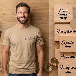 May include: A man wearing a tan t-shirt with "Super Papa" and a heart graphic. Wooden blocks with phrases like "Papa d'amour," "Dad of two," and "Daddy cool" are displayed. A comb, compass, and decorative elements are also visible.