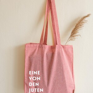 May include: A red tote bag with long straps hangs from a wooden branch against a cream-colored wall. The bag has white text that reads "EINE VON DEN JUTEN". A dried plant stem is tucked inside the bag.
