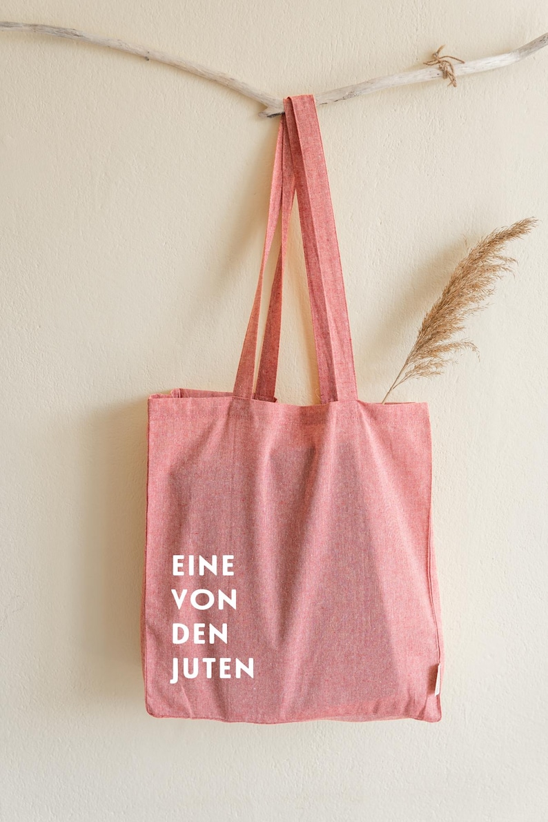 May include: A red tote bag with long straps hangs from a wooden branch against a cream-colored wall. The bag has white text that reads "EINE VON DEN JUTEN". A dried plant stem is tucked inside the bag.