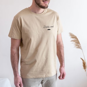 May include: A beige t-shirt with the text "Daddy cool" and a small black mustache graphic. The shirt is a crew neck style and appears to be made of a soft material. The model is wearing beige pants.