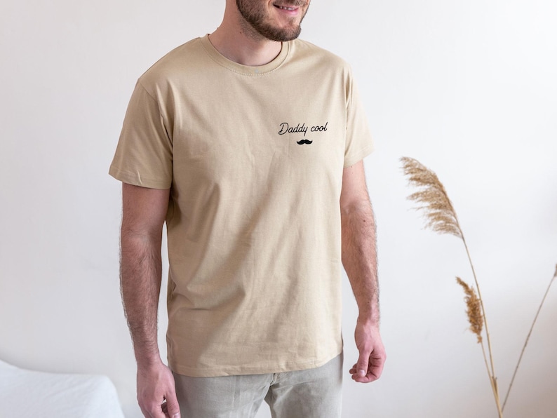 May include: A beige t-shirt with the text "Daddy cool" and a small black mustache graphic. The shirt is a crew neck style and appears to be made of a soft material. The model is wearing beige pants.
