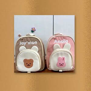 May include: Two small backpacks with white front pockets and animal-themed patches. The left backpack is tan with the name Jay'vion and a bear patch. The right backpack is pink with the name Anaia and a pink rabbit patch, both featuring ears.