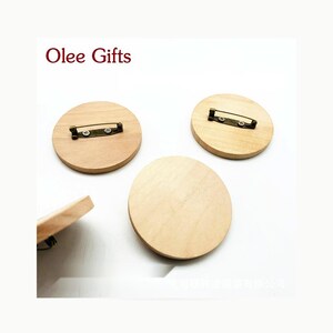 Puede incluir: Tres piezas circulares de madera de color claro para broches con superficies lisas. Dos de los discos de madera cuentan con cierres de alfiler de metal en tono bronce fijados con tornillos pequeños. El texto Olee Gifts aparece en rojo.