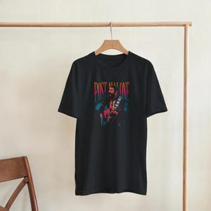 May include: Black t-shirt featuring a colorful graphic of a musician playing a guitar. The text "POST MALONE" is printed above the graphic. The shirt is hanging on a wooden hanger.