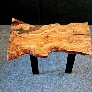 Peut inclure: Une table en bois à bords naturels de forme organique. Le plateau est de couleur marron chaud avec des motifs de grain de bois complexes. La table est soutenue par deux pieds noirs rectangulaires.