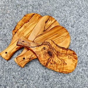 Op de afbeelding: Een verzameling van drie olijfhouten serveerplanken. Elke plank heeft een uniek houtnerfpatroon en een handvat met een gat om op te hangen. De ovale planken hebben een natuurlijke, rustieke uitstraling. Het hout heeft een warme, lichtbruine kleur.