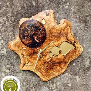 May include: A handcrafted wooden wall clock with a natural edge. The clock features a round face with a tree design and gold-colored hands. The clock is made of olive wood and has a unique, organic shape. The clock measures approximately 12 inches in diameter.