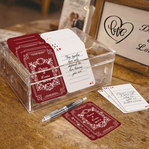 May include: A clear acrylic box holds playing cards with burgundy backs and white fronts. The burgundy cards feature ornate white designs and the names "Darwin & Louisa." White cards have handwritten text. A pen rests nearby. Wedding theme.