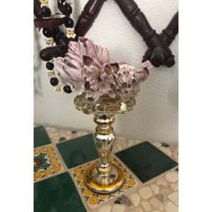 May include: A decorative glass pedestal with a silver finish and gold accents, topped with a cluster of pink and white barnacles. The pedestal sits on a mosaic tile surface, with a beaded necklace and wooden wall decor in the background.