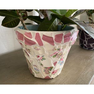 May include: A mosaic flower pot with a light blue interior. The pot is made of broken pieces of white ceramic with pink and floral patterns. The pot is filled with a green plant.