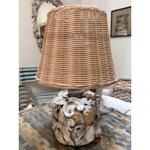 May include: A table lamp featuring a cylindrical base covered in overlapping natural oyster shells in shades of gray, brown, and white. The lamp is topped with a tapered, light tan woven wicker lampshade with a visible horizontal weave pattern.