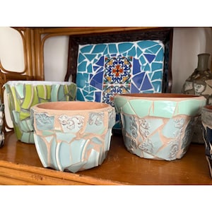 May include: A collection of decorative ceramic flower pots with a mosaic design. The pots are various sizes and colors, including light blue, green, and white. The mosaic pattern is made of broken pieces of ceramic tiles.