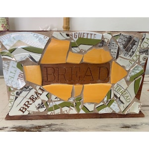 May include: A wooden bread box with a mosaic design made from broken pieces of ceramic tiles. The tiles are in shades of yellow, green, and white. The word "BREAD" is carved into the front of the box.
