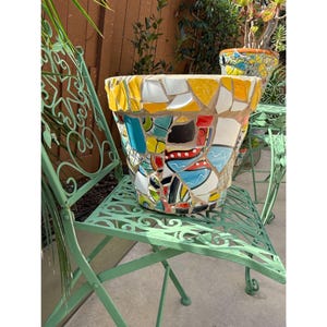 May include: A large, mosaic-tiled planter pot with a wide rim, resting on a green metal chair. The pot features a colorful design of yellow, white, blue, and black tiles. Another mosaic pot is visible in the background.