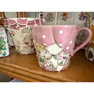 Pode incluir: Uma chávena de chá rosa em mosaico com bolinhas brancas e uma pega. A chávena é decorada com peças de cerâmica com motivos florais. Um vaso de mosaico a condizer está no fundo, juntamente com outras obras de arte em mosaico.