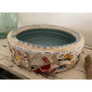 May include: A round mosaic planter with a blue interior. The planter is made of broken ceramic tiles in various colors, including white, red, yellow, and blue. The tiles are arranged in a random pattern.