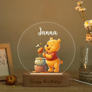 Può includere: Una luce notturna decorativa a tema Winnie the Pooh, con il nome "Janna" su un pannello in acrilico trasparente. La luce poggia su una base in legno incisa con "Happy Birthday". L'illustrazione mostra Pooh con un barattolo di miele. Un vaso di fiori è sullo sfondo.