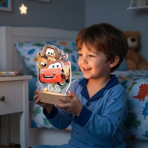 Może przedstawiać: Podświetlana lampka nocna z postaciami z filmu Auta, w tym Zygzakiem McQueenem i Złomkiem. Światło jest zamontowane na drewnianej podstawie i ma imię "Caleo". Tło przedstawia sypialnię z łóżkiem i stolikiem nocnym.