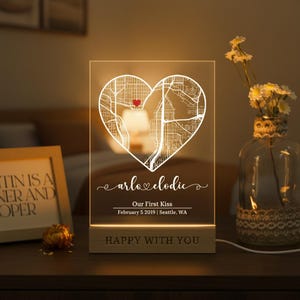 May include: A personalized LED night light with a heart-shaped map design. The light displays the text "arlo & clodie", "Our First Kiss", and the date and location. The base of the light reads "HAPPY WITH YOU".